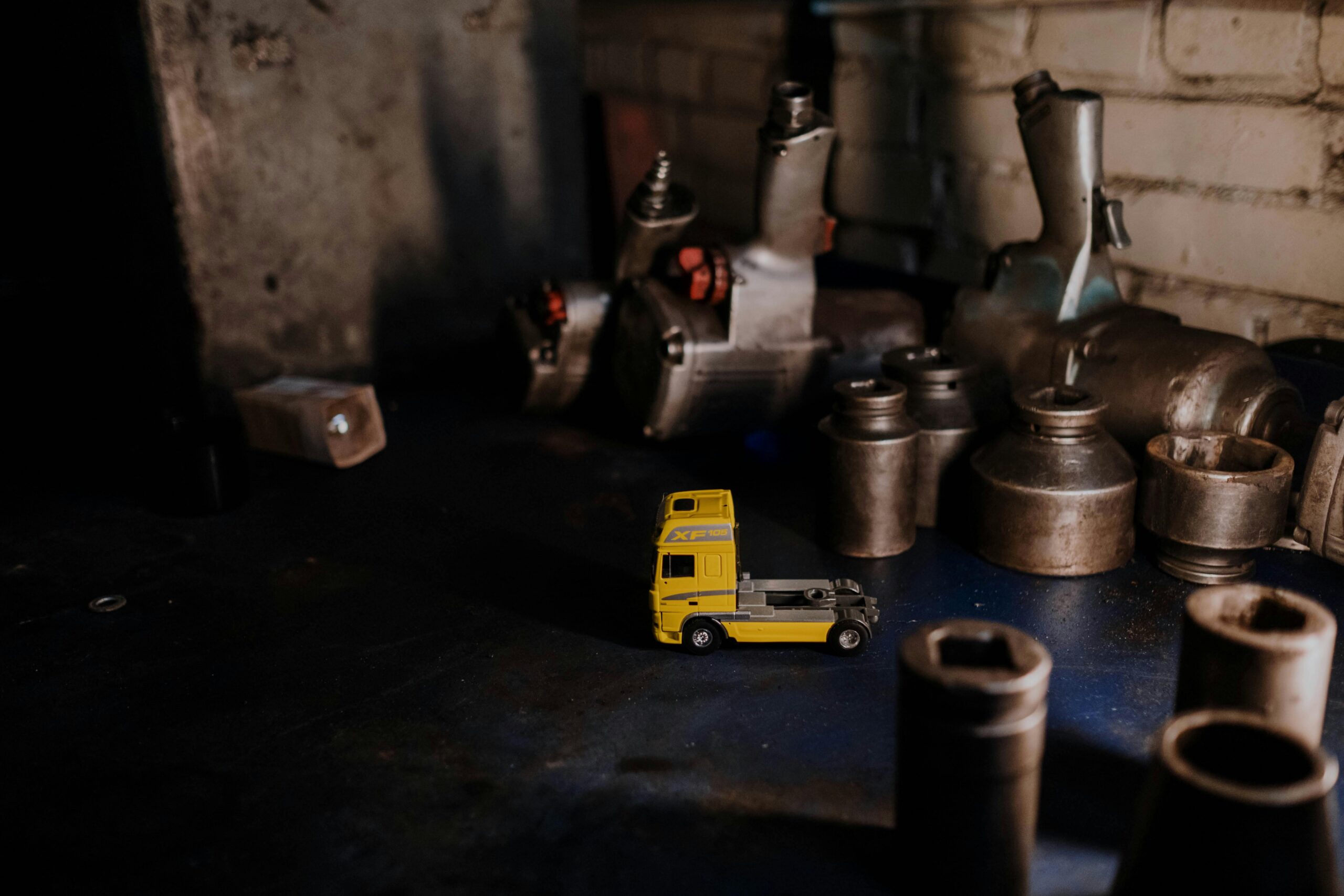 A miniature yellow toy truck in an industrial workshop surrounded by metal parts.